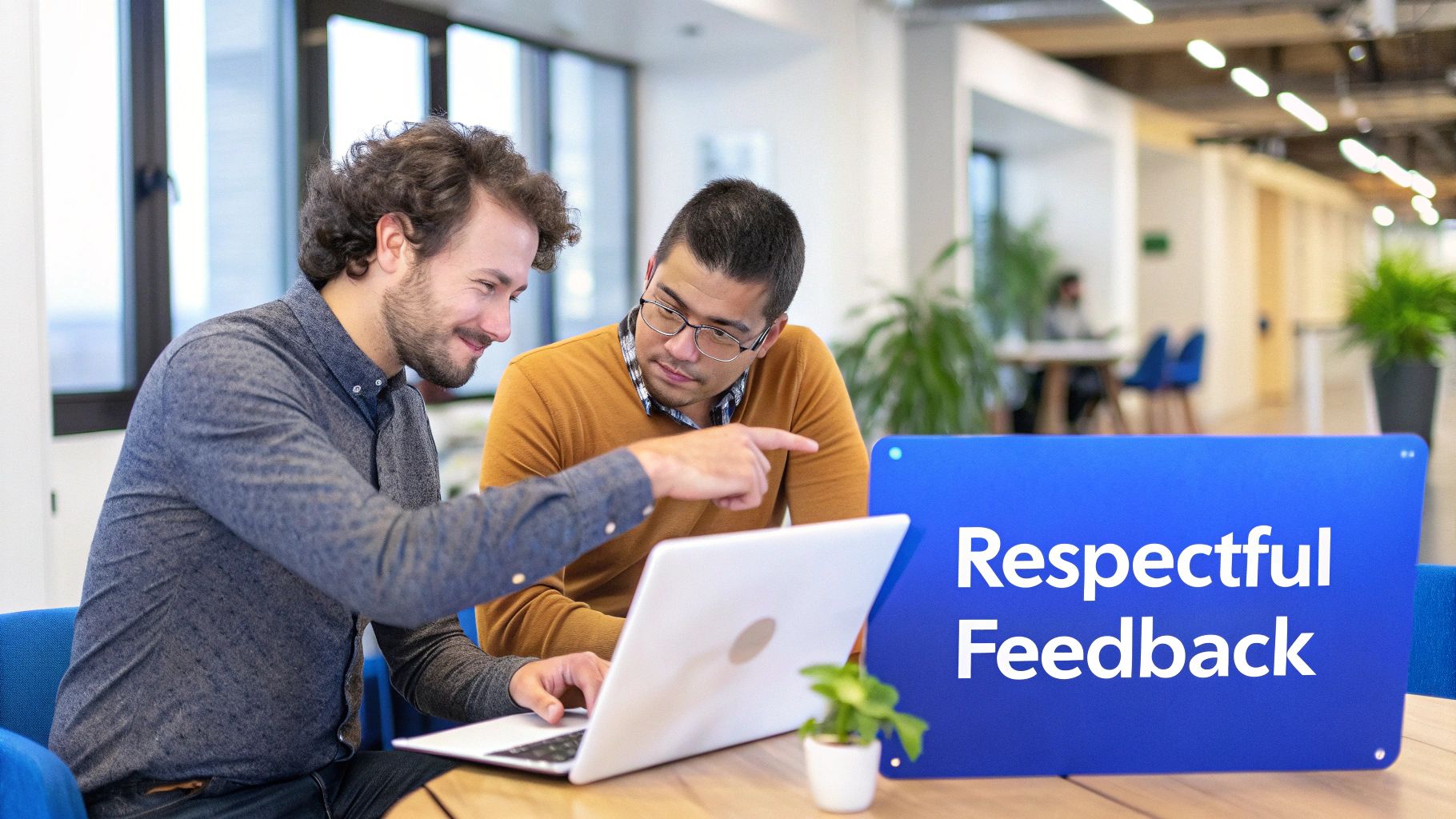 Two men collaborating on a laptop in an office, with a 'Respectful Feedback' sign on the table.